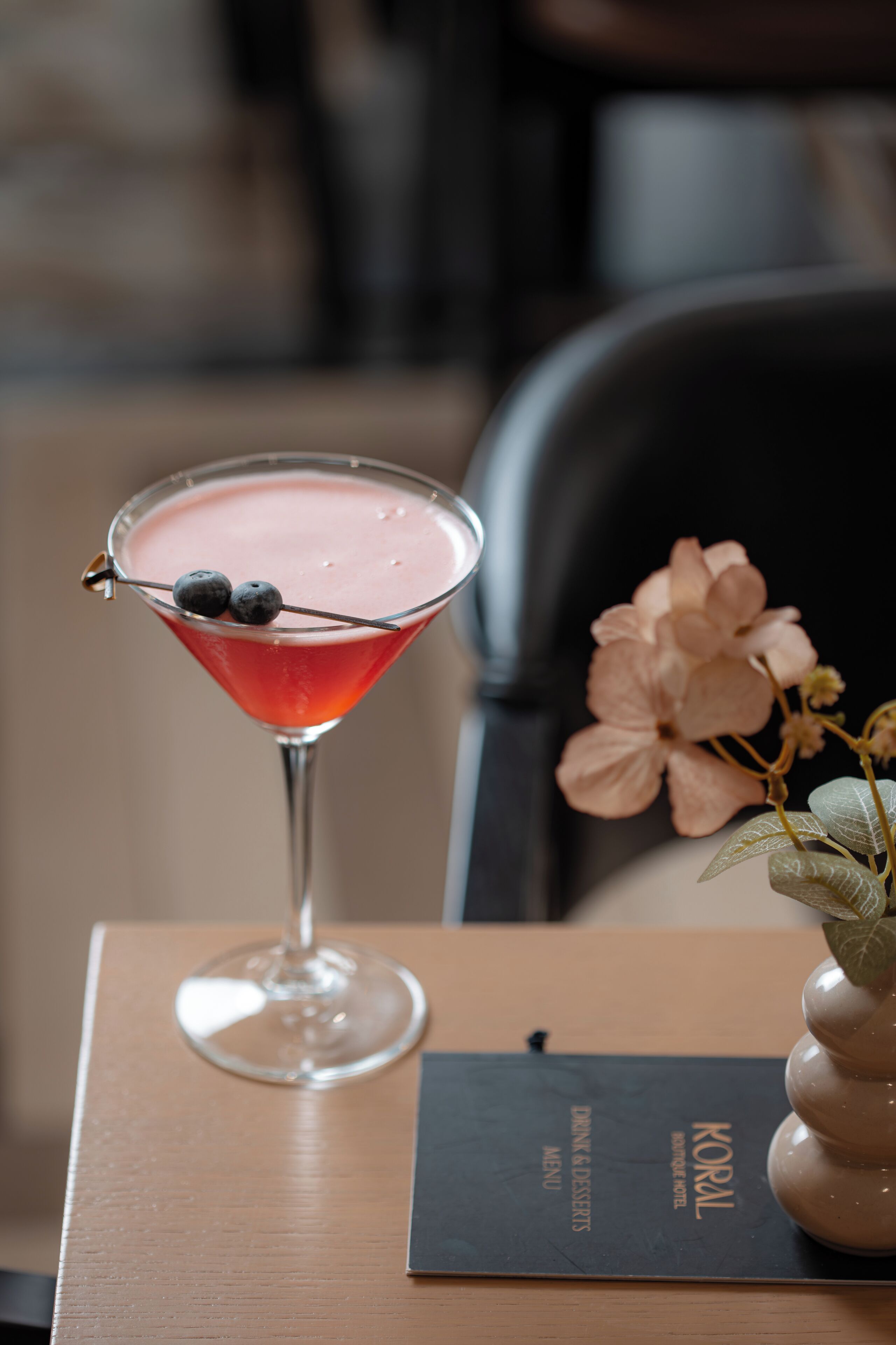 A martini glass with a pink drink, blueberries, and a flower on a table with a menu.