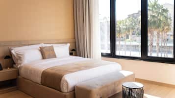 Habitación de hotel con una cama grande, una mesita de noche con lámpara, una mesita redonda y vistas a palmeras y edificios.