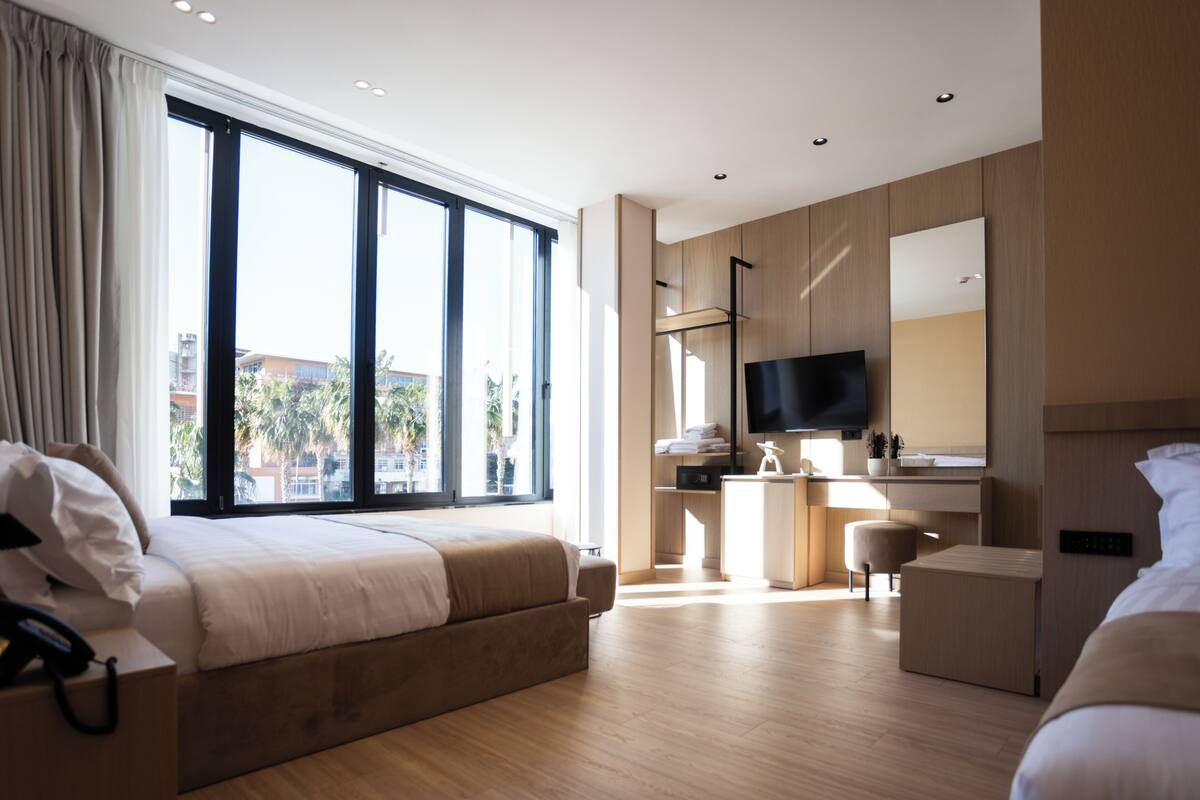 A modern hotel room with a large window, a bed, a desk, and a TV.