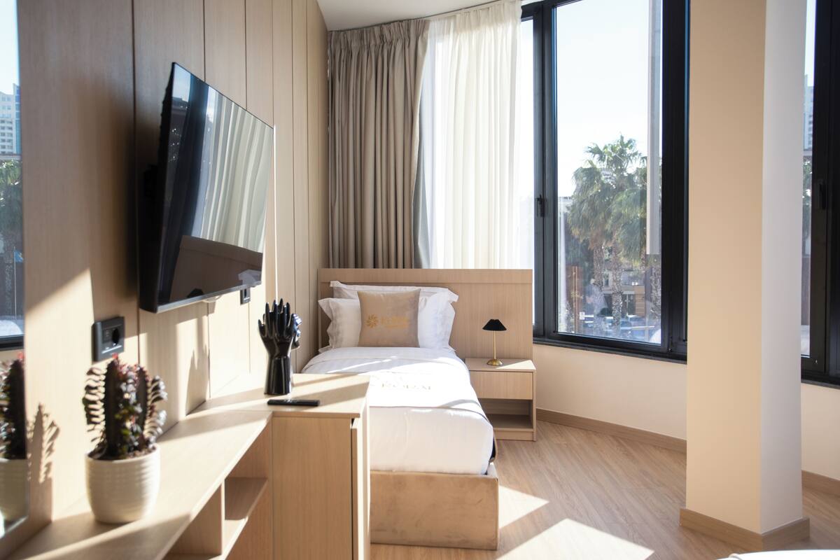 A modern hotel room with a bed, bedside table, TV, and large windows.