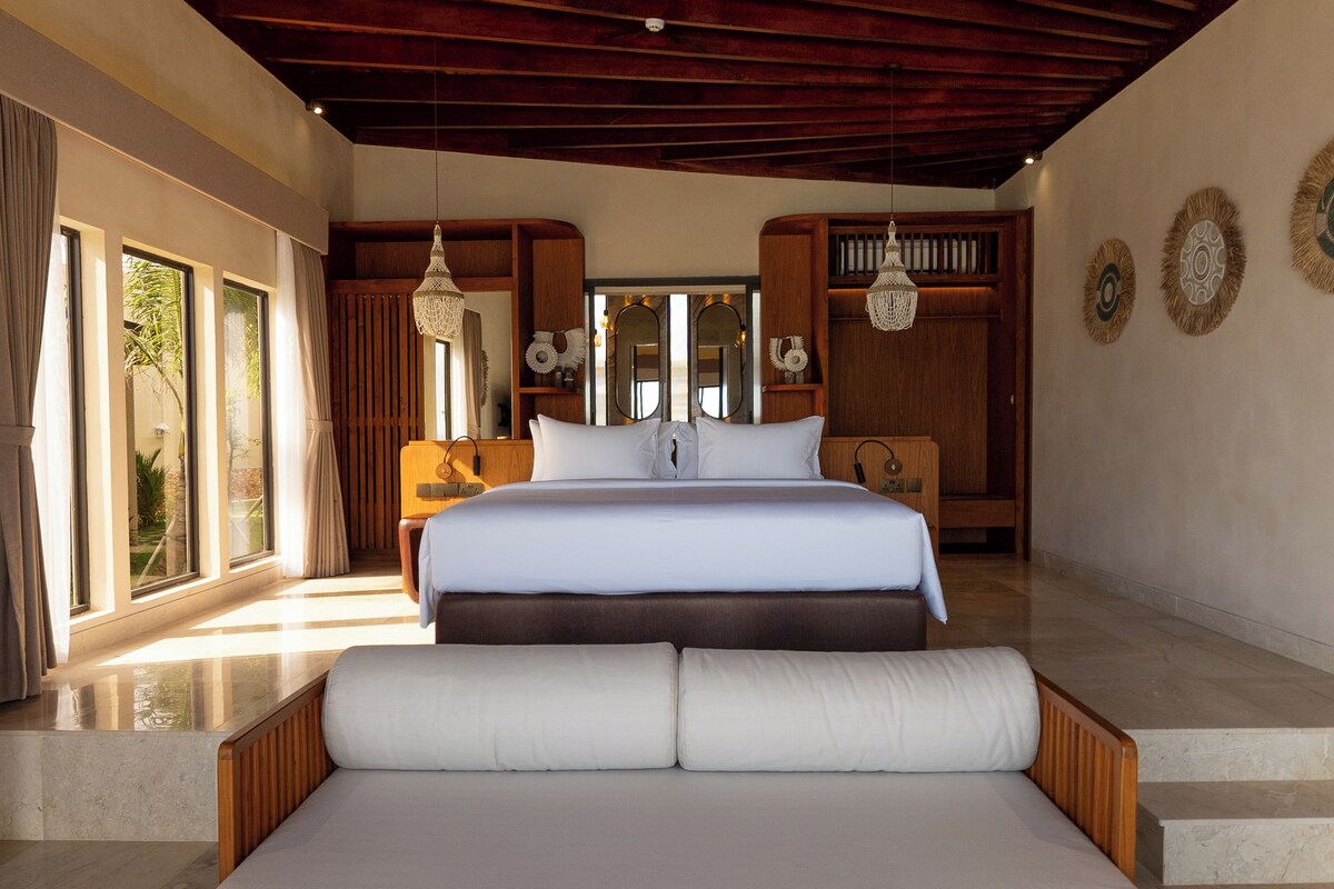 A spacious bedroom with a large bed, wooden ceiling beams, and a view of the outdoors through large windows.