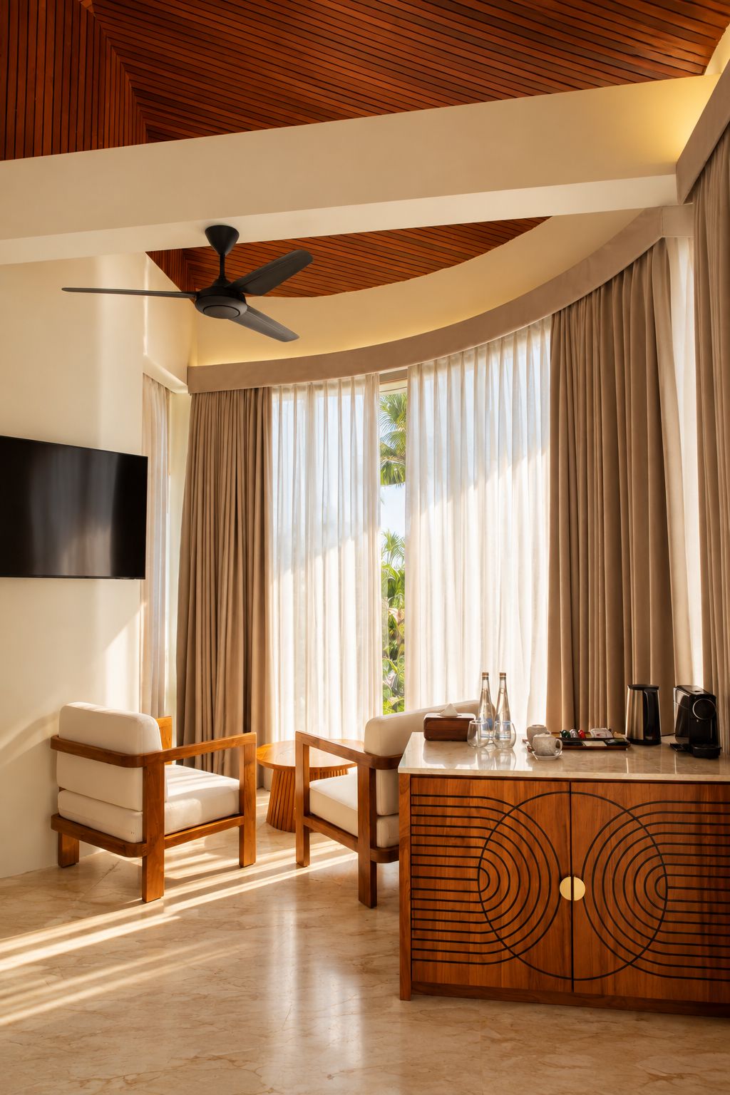 A modern living room with a ceiling fan, a TV mounted on the wall, a wooden cabinet with decorative patterns, and a large window with curtains.