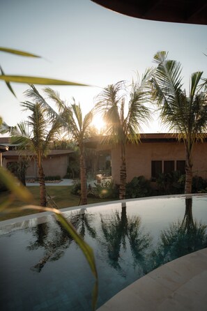 A swimming pool with palm trees, a building with a curved roof, and a clear sky. - VANARA Resort & Spa (Pecatu)