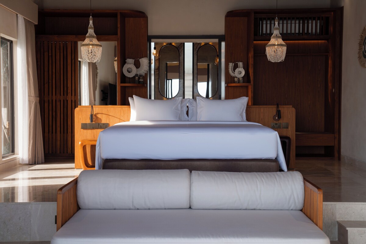 A modern bedroom with a large bed, wooden headboard, bedside tables, and two hanging light fixtures.