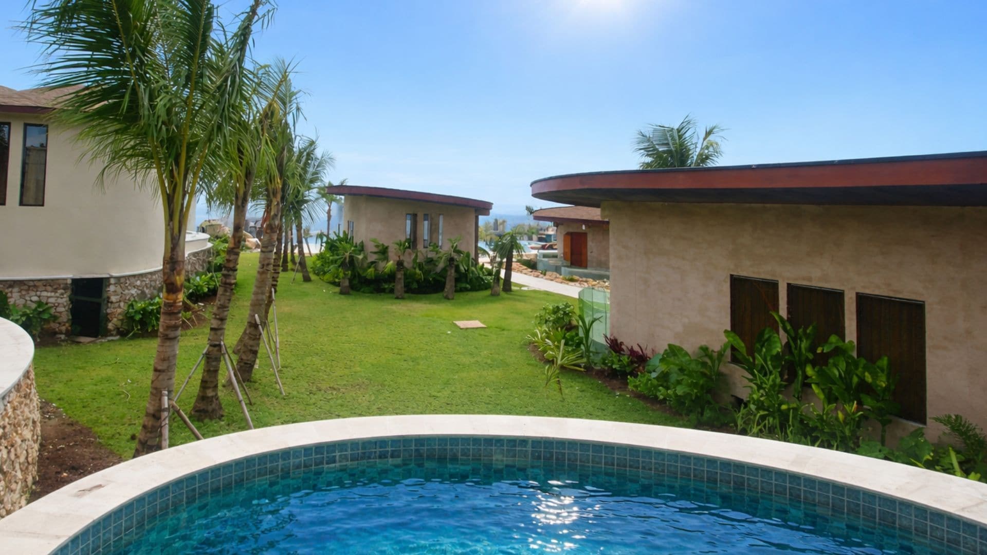 A pool area with a clear view of the ocean, surrounded by palm trees and a well-maintained lawn.
