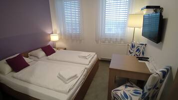 Une chambre d’hôtel avec deux lits, un bureau, une chaise, une télévision et deux fenêtres équipées de stores.