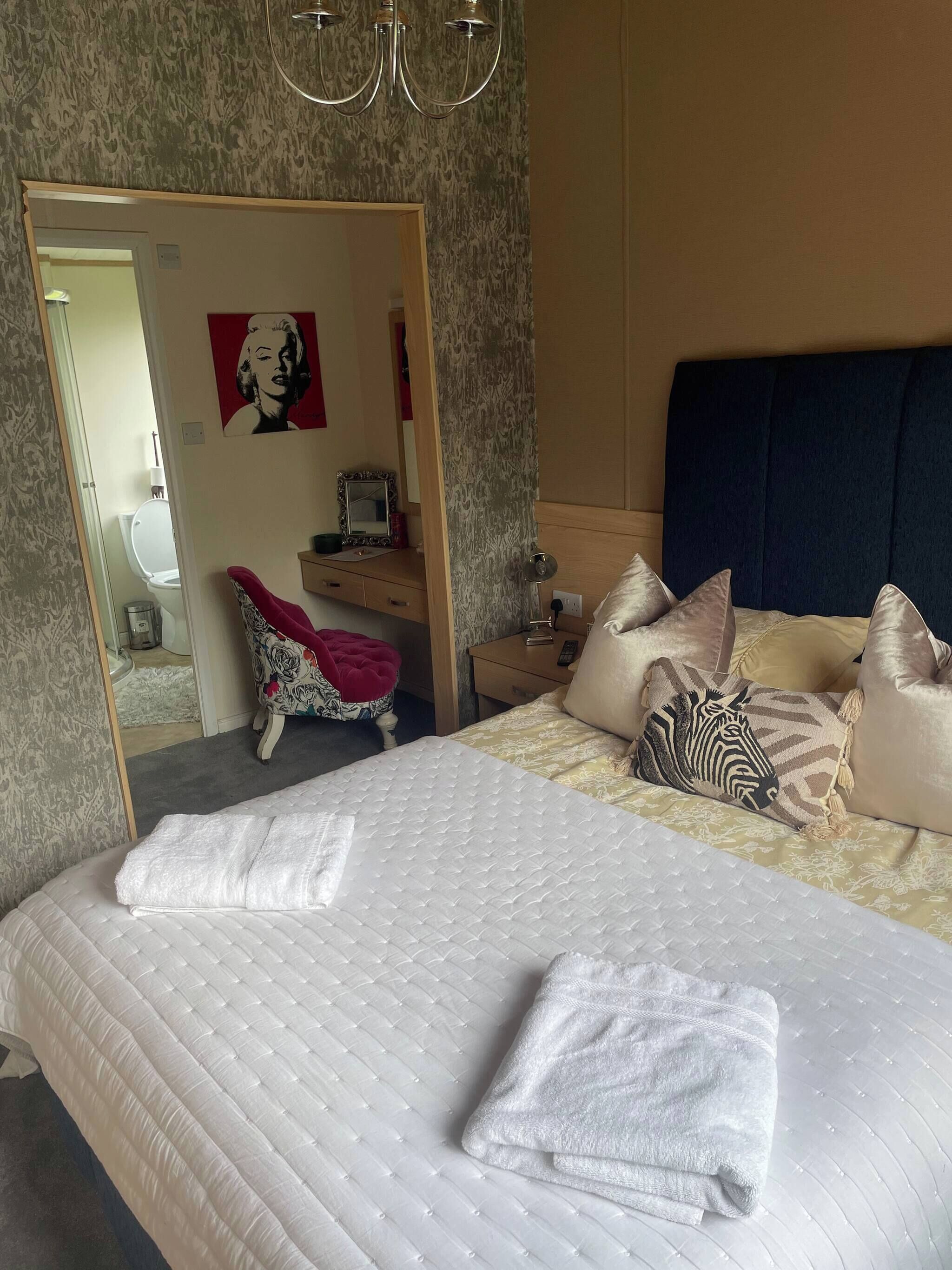 A bedroom with a bed, pillows, a bedside table, a mirror, and a door leading to a bathroom.