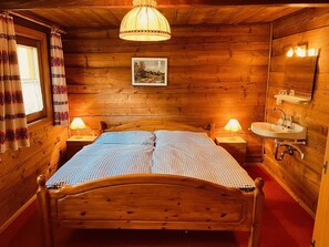 Un dormitorio de cabaña de madera con cama, lámparas de noche, lavamanos empotrado y un cuadro enmarcado en la pared.