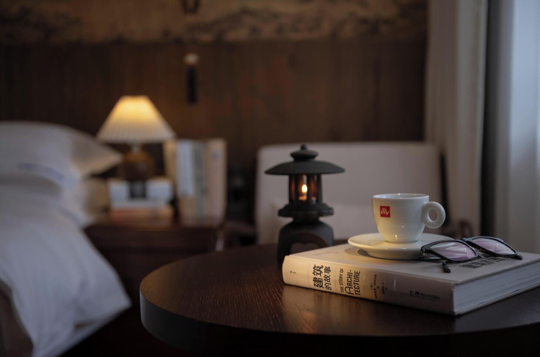 A round table with a book, a cup, and a pair of glasses. A lantern is in the background.