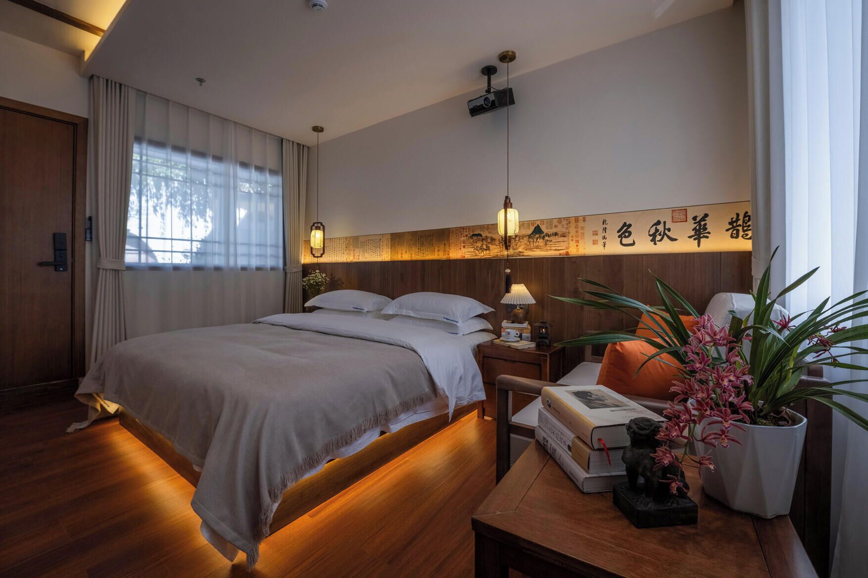 Quarto de hotel com cama, mesa de cabeceira com livros e uma luminária, uma planta em vaso e parede com caligrafia.