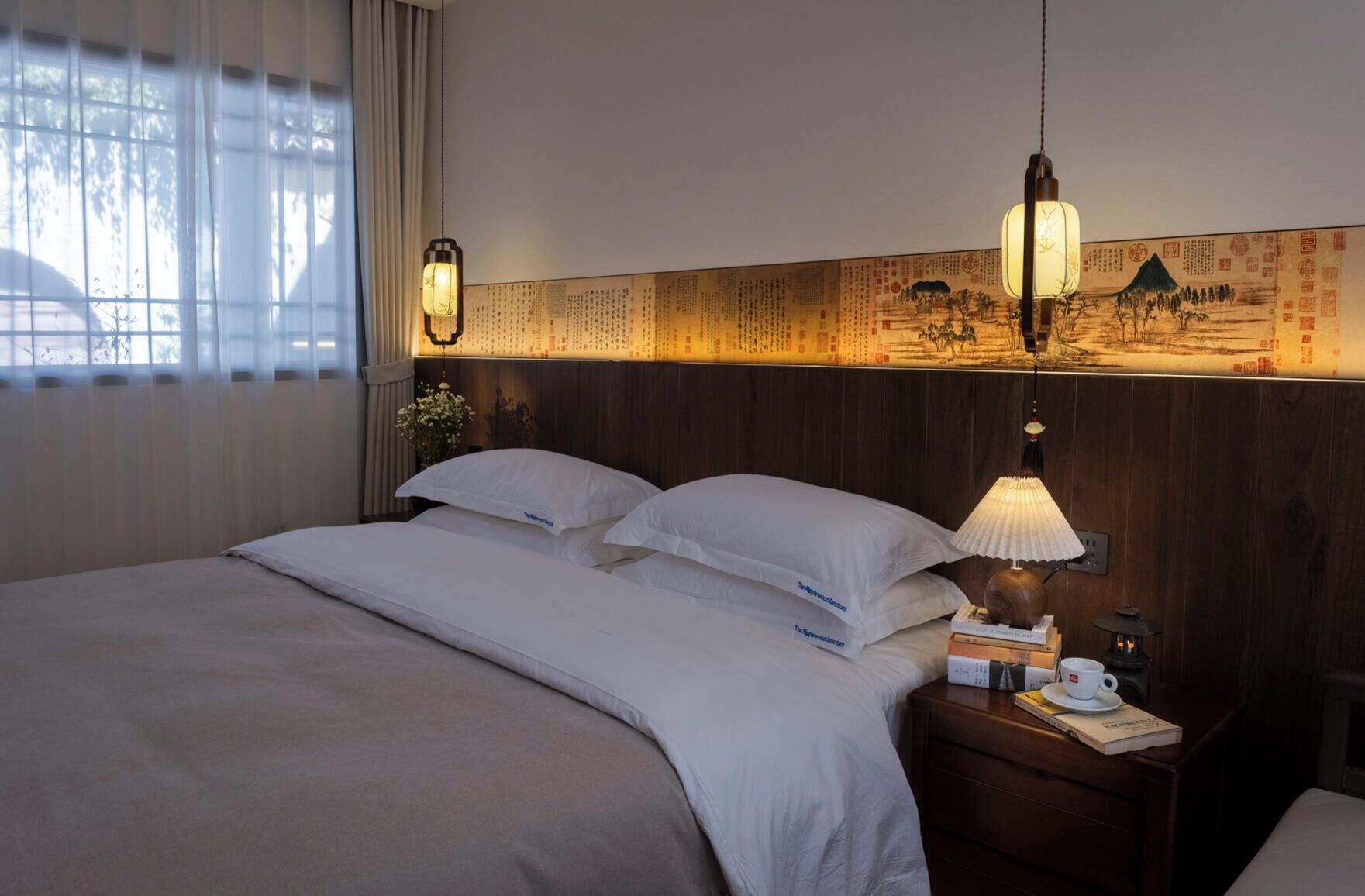 Une chambre d’hôtel avec un lit, une table de chevet avec une lampe, une petite table de nuit avec un livre et une tasse, et un mur orné d’œuvres d’art traditionnelles.