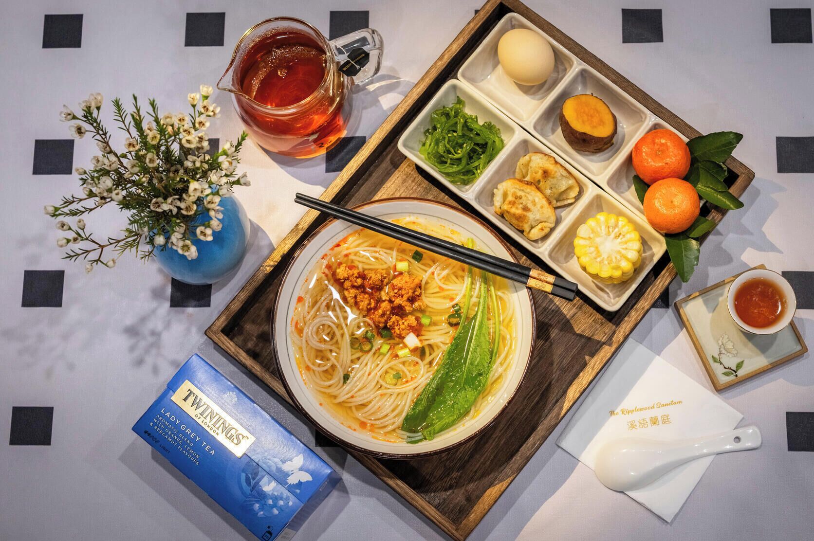 Una mesa con un tazón de fideos, una jarra de té, una caja de té y una bandeja con diferentes alimentos.