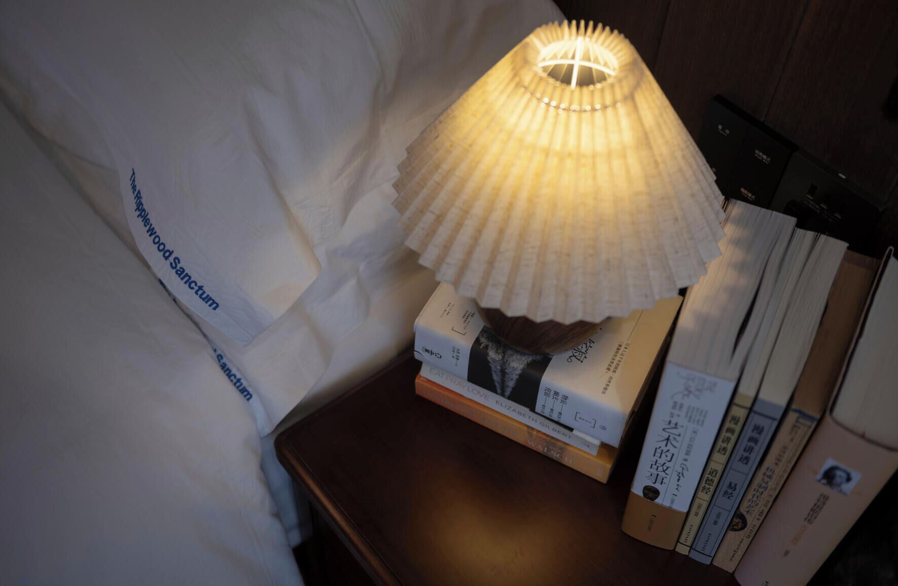 A bedside table with a lamp, books, and a pillow with text.