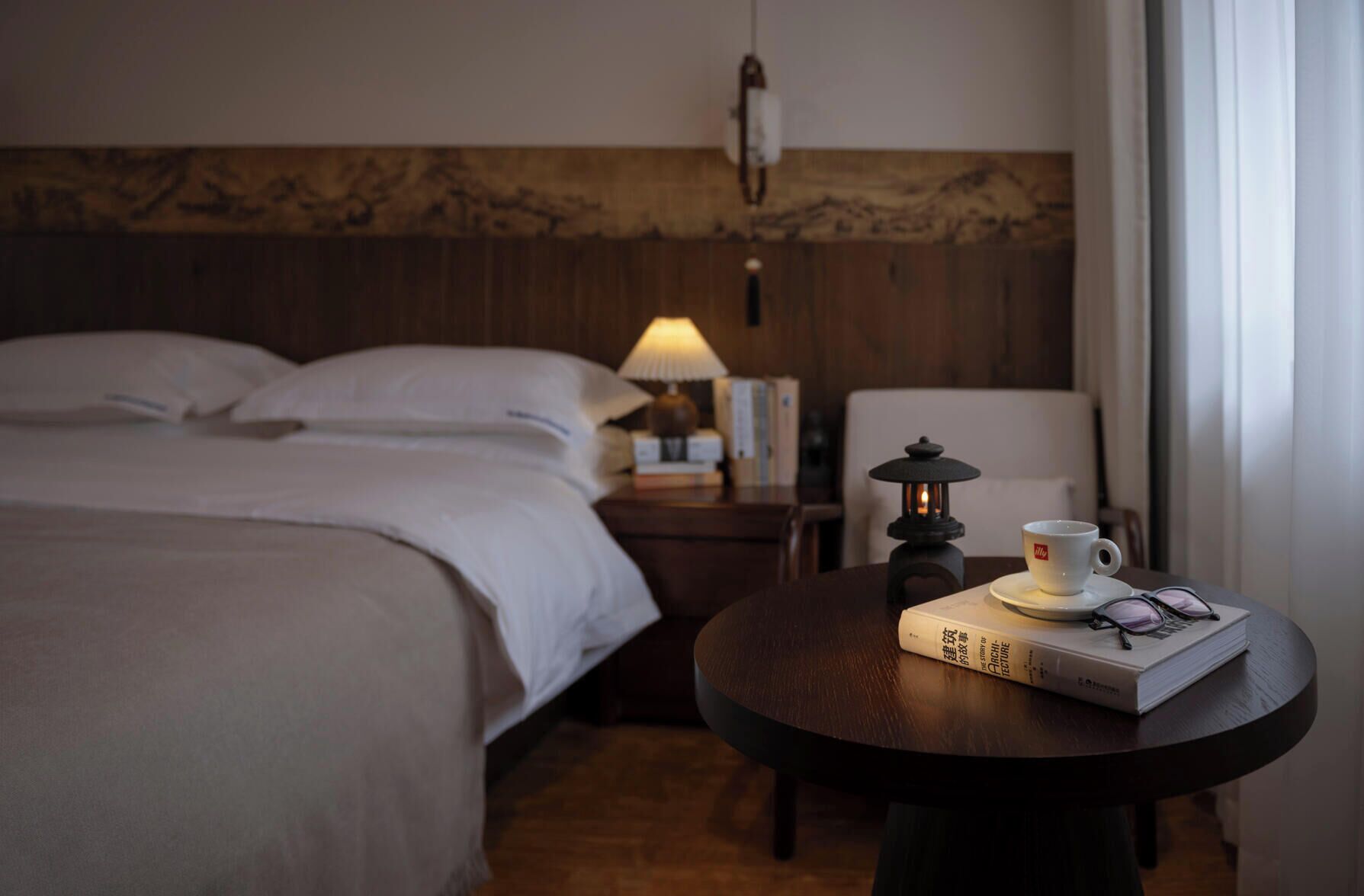 A hotel room with a bed, a nightstand with a lamp, a table with a book, a cup, and glasses.
