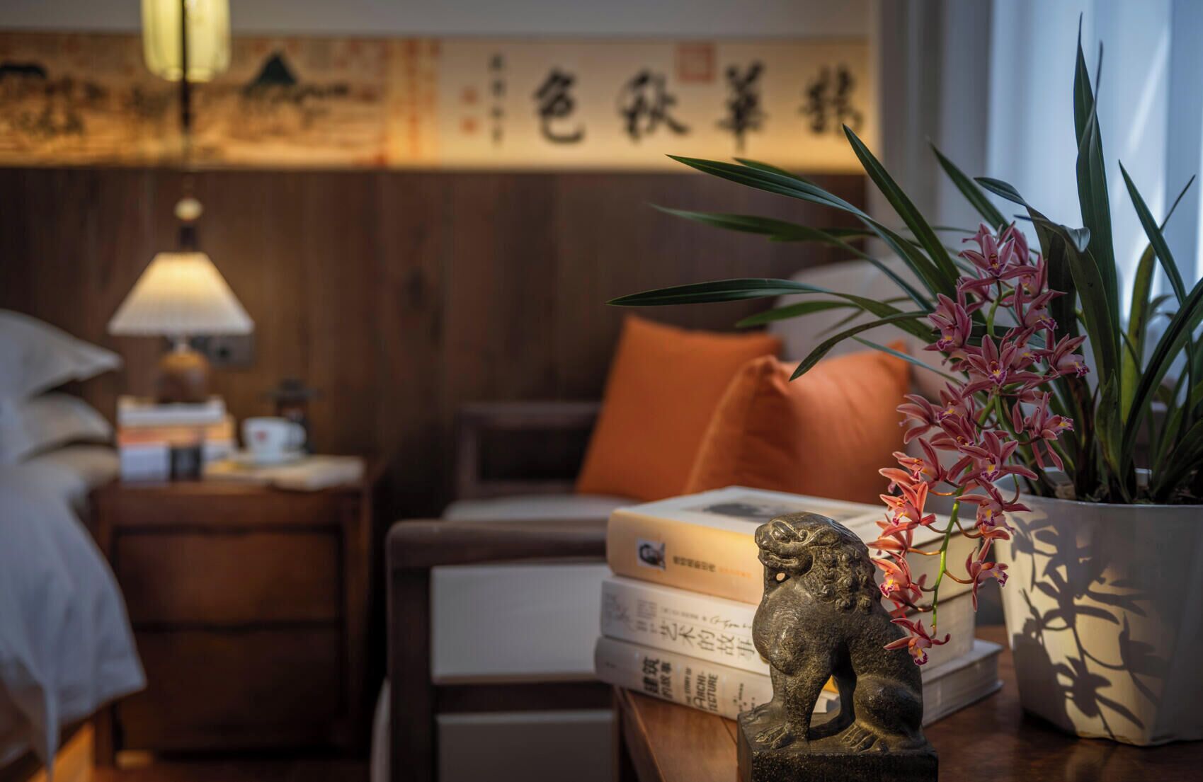 Une chambre à coucher bien rangée, avec un lit, une table de chevet, une lampe, des livres, une statue décorative et une plante en pot.
