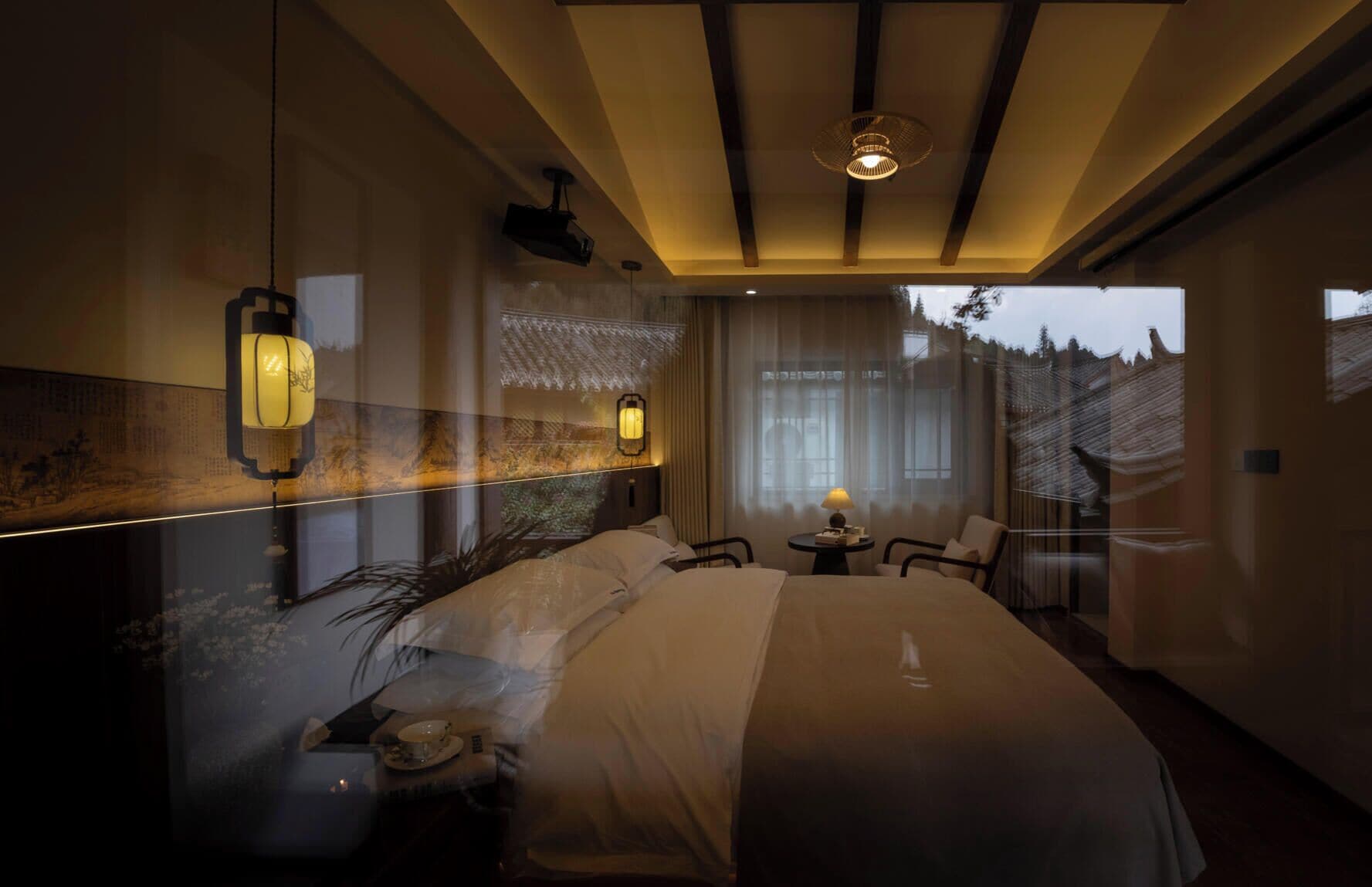 Ein modernes Hotelzimmer mit einem großen Bett, einem kleinen Tisch mit Lampe und Blick ins Freie durch ein Fenster.