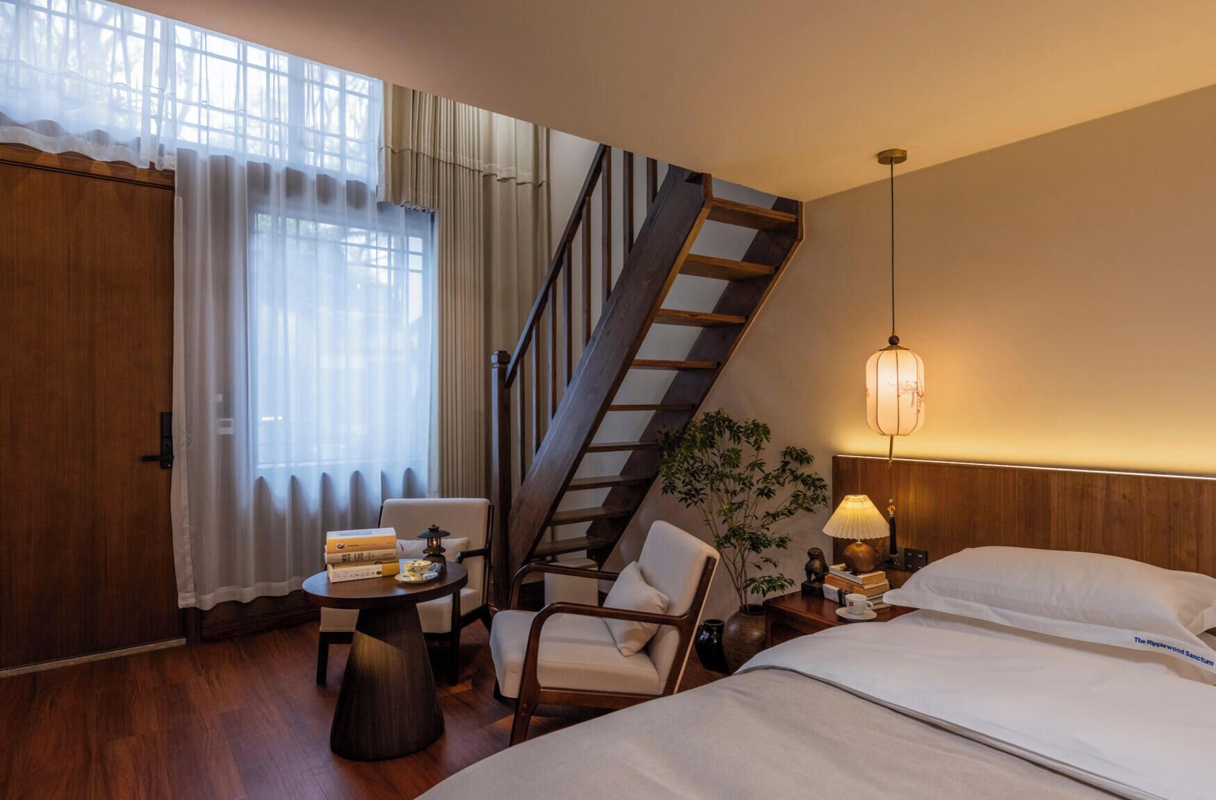 A hotel room with a bed, a chair, a small table with books and a lamp, a staircase, and a window with curtains.
