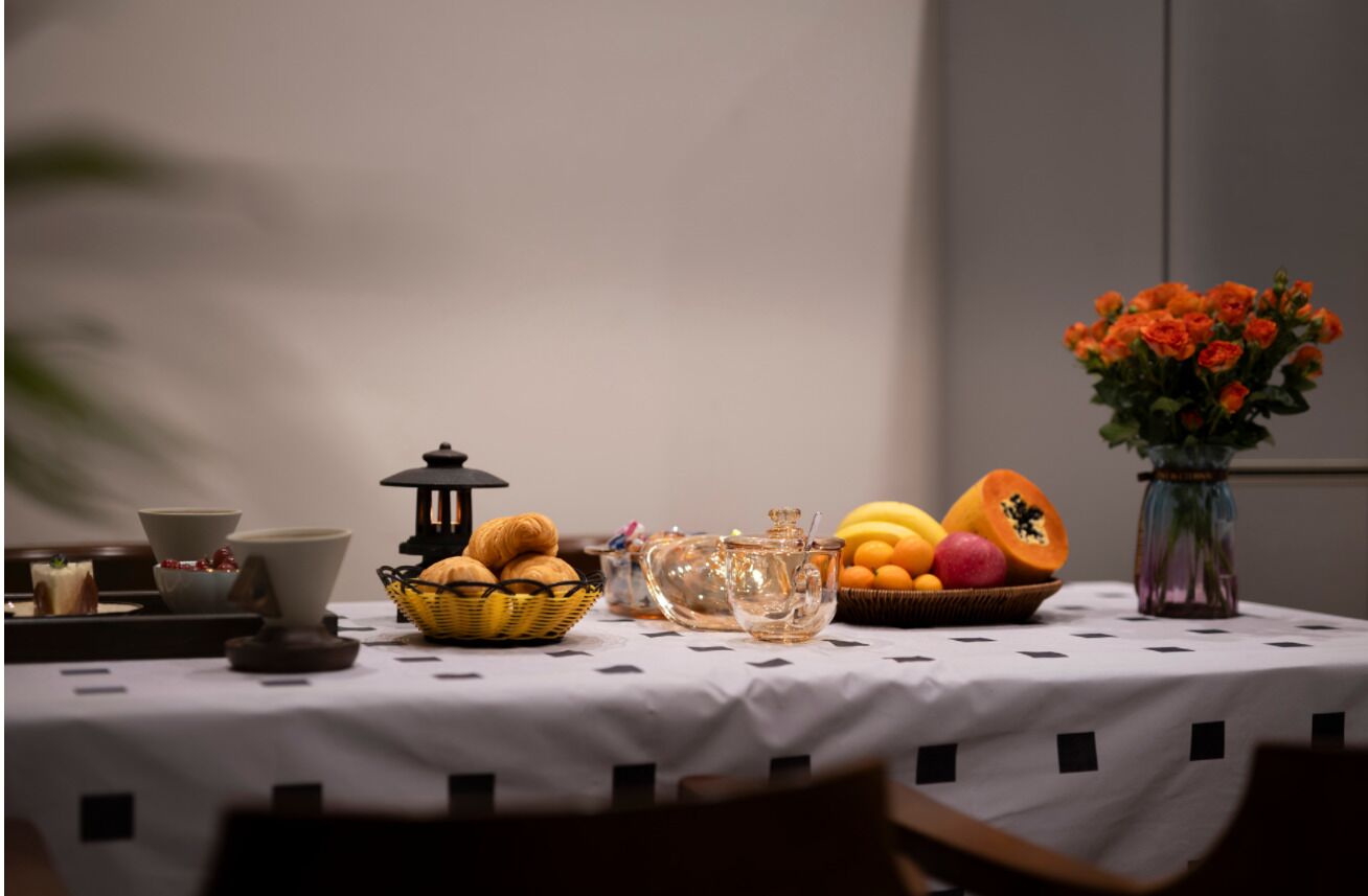 Mesa com toalha branca, cesta de pão, jarra de vidro, tigela de frutas e vaso com flores.