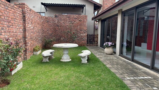A courtyard with a brick wall, a small garden, a round table with four chairs, and a glass building entrance.