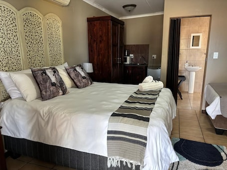 A bedroom with a large bed, a wooden wardrobe, a small kitchenette, and a bathroom with a sink.