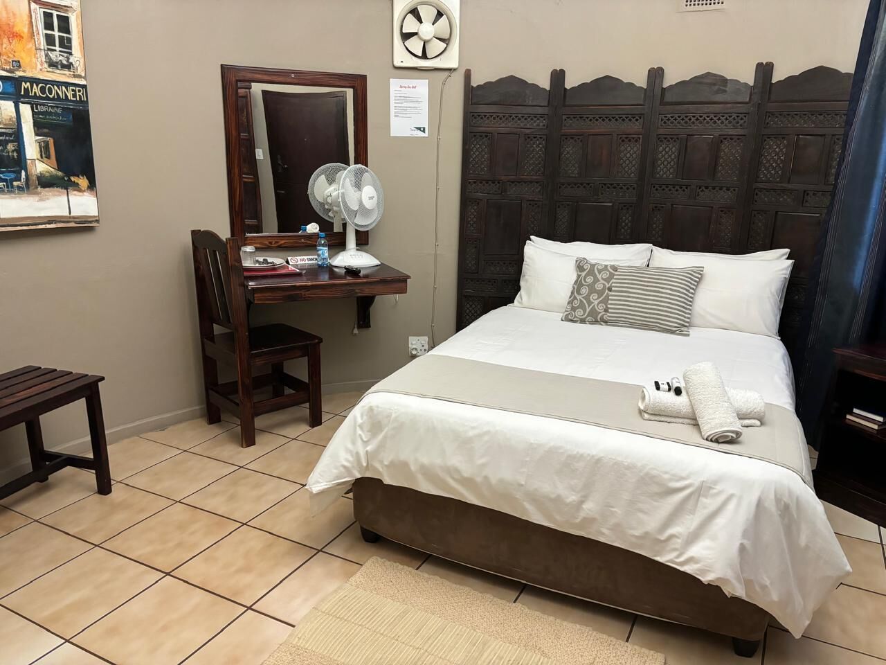 Ein Hotelzimmer mit Bett, Nachttisch, Sessel, Schreibtisch mit Spiegel und einem Bild an der Wand.