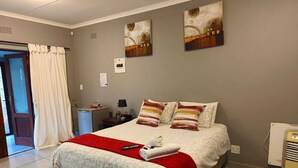 A neatly made bed with a red blanket, a remote control, and a towel on the bed. There are two paintings on the wall, a nightstand with a lamp, and a heater on the floor.