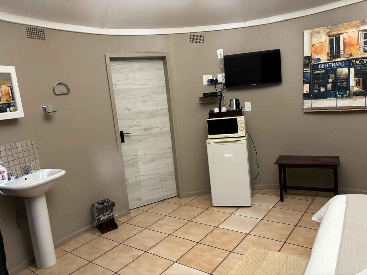 A compact room with a white sink, a small refrigerator, a flat-screen TV mounted on the wall, a door, and a small table.