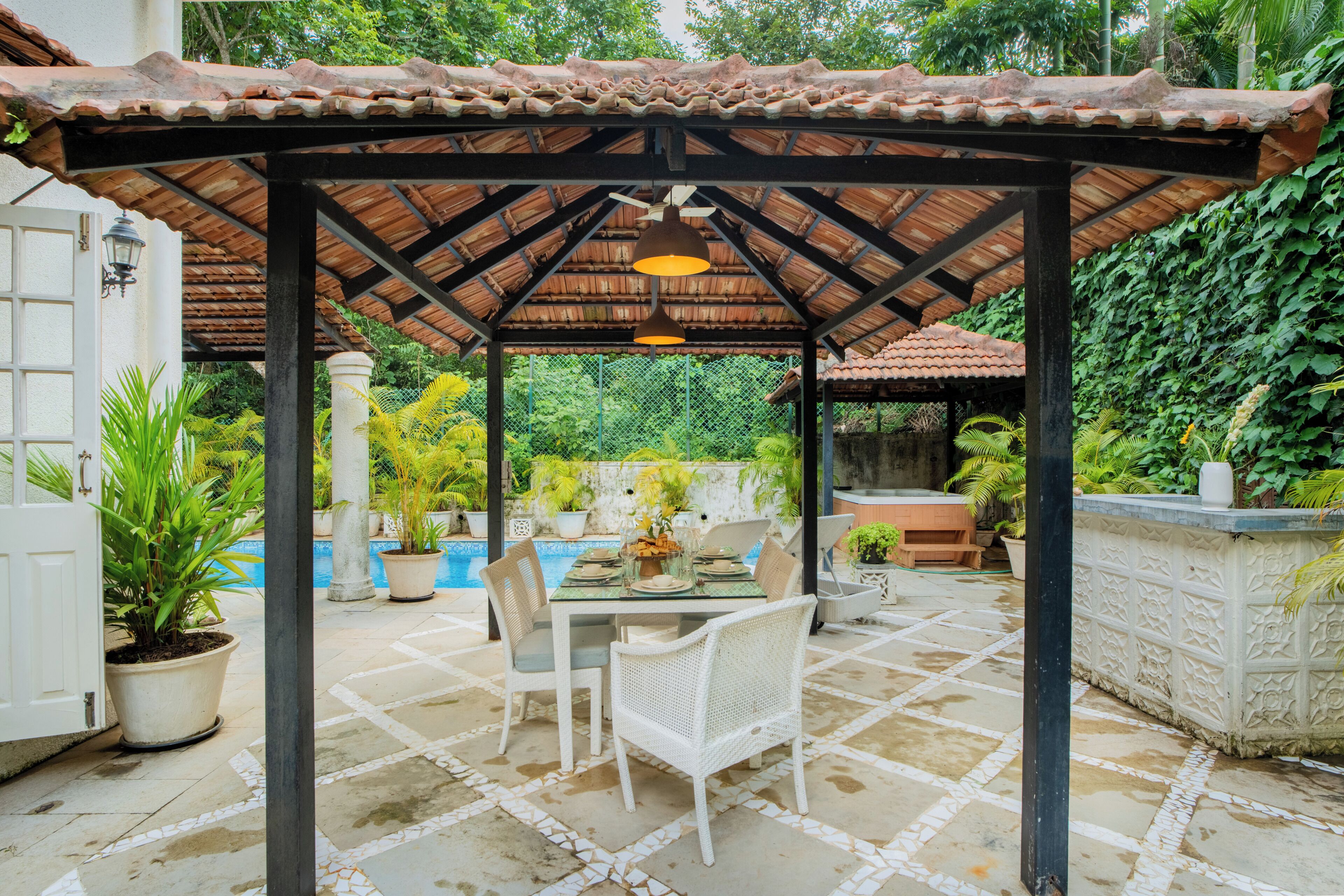 A covered outdoor seating area with a table and chairs, surrounded by potted plants and a swimming pool.
