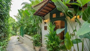A narrow pathway flanked by lush greenery and a building with a tiled roof and green accents. - ELIVAAS Monforte Ayada 3 BHK Villa (Assagao)