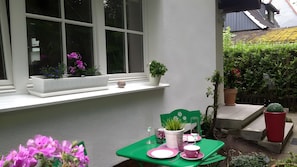 Ein kleiner Sitzbereich im Freien mit Tisch und Stühlen, ein Fenster mit Blumentöpfen und Blick auf einen Garten.
