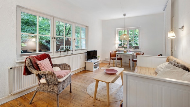 Une pièce confortable avec un fauteuil en osier, une petite table, un canapé et un téléviseur. De grandes fenêtres offrent une vue sur la verdure environnante.