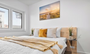 A bedroom with a bed, pillows, a blanket, a painting, a window, and a bedside table with a plant. - Vacation apartment Waterkant Büsum directly on the beach with sea view over the harbor (Büsum)