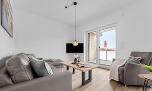 A modern living room with a sofa, a coffee table, a TV, and a balcony with a view of a tower. - Vacation apartment Waterkant Büsum directly on the beach with sea view over the harbor (Büsum)