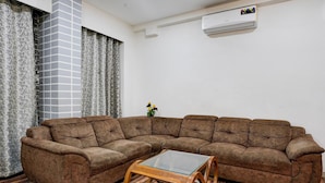 Ein Wohnzimmer mit einem Ecksofa, einem Couchtisch mit Glasplatte, gemusterten Vorhängen und einer Klimaanlage.