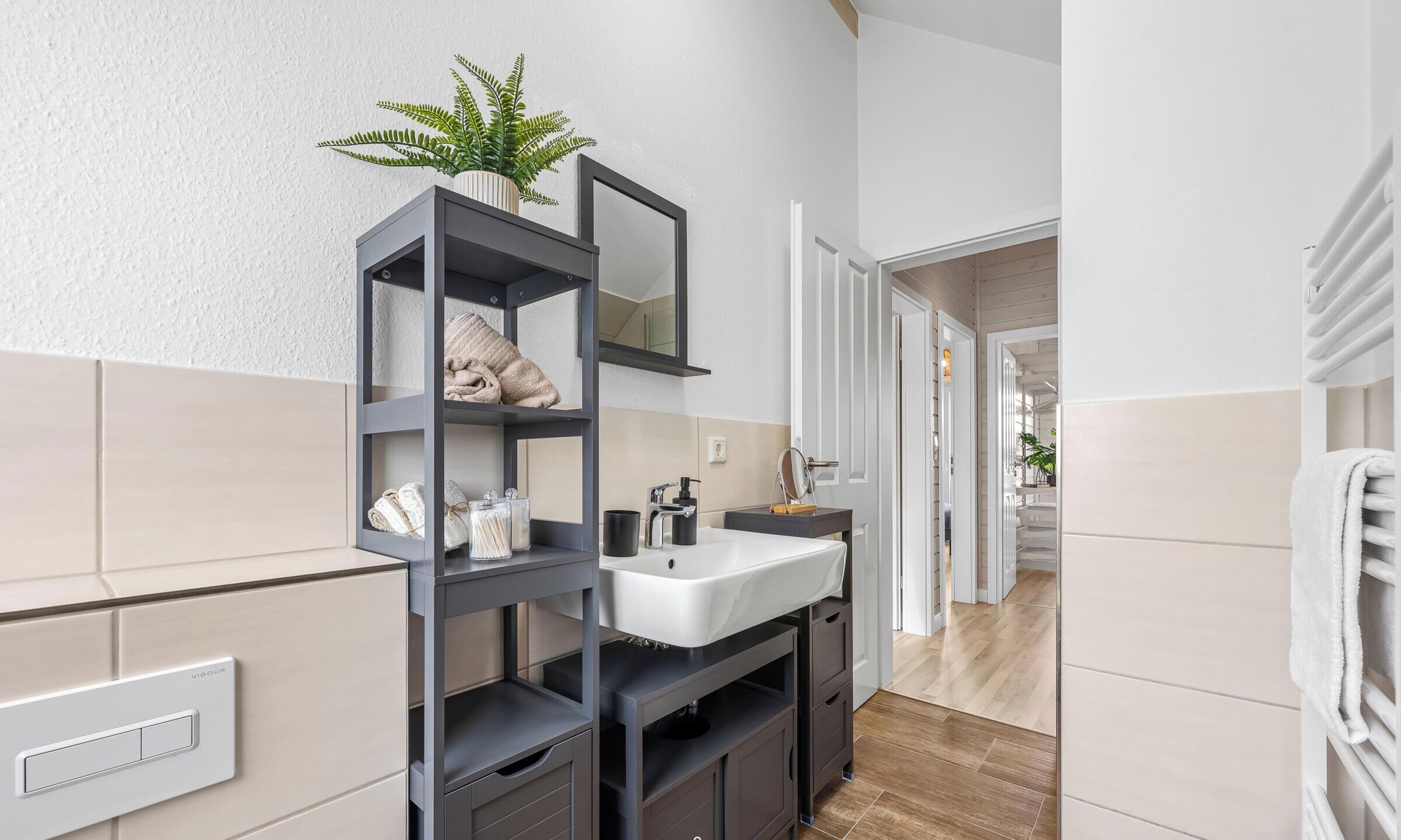 A modern bathroom with a grey vanity, white sink, and a mirror. There is a potted plant on the vanity and a towel rack to the right.