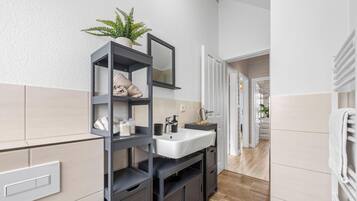 A modern bathroom with a grey vanity, white sink, and a mirror. There is a potted plant on the vanity and a towel rack to the right.