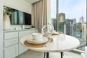 A modern dining area with a round table, white plates, and cutlery. A large window offers a city view. - Stunning Canal View, Studio with Balcony - Peninsula Three, Business Bay (Dubai)