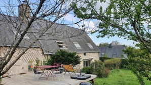 Ein Hinterhof mit einer Holzterrasse, Gartenmöbeln, einem Grill und einem Haus mit einem geneigten Dach und Steinverkleidung.