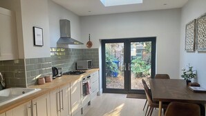 A modern kitchen with a gas stove, microwave, and sink, a dining area with a table and chairs, and a sliding glass door leading to a garden. - Delightful 2 Bedroom House Central Windsor. Walk to Castle, River! (Windsor)