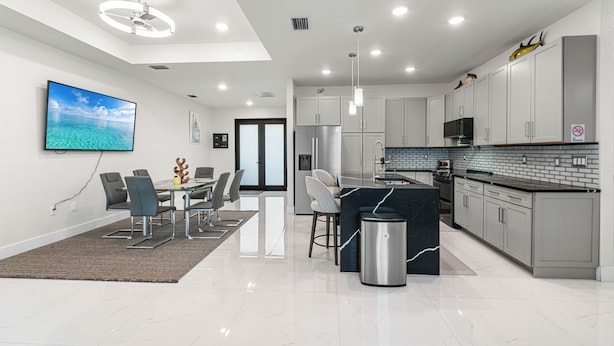 A modern kitchen with a dining area, featuring a large island, stainless steel appliances, and a flat-screen TV mounted on the wall. - Nick House (Lehigh Acres)
