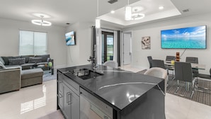 A modern kitchen with a black countertop, a dining area with a glass table, and a living room with a grey sofa. - Nick House (Lehigh Acres)