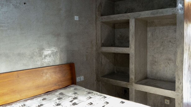 A room with a wooden headboard, a bed with patterned bedding, and a concrete shelving unit against the wall. - Monkey Palms (Gili Air)