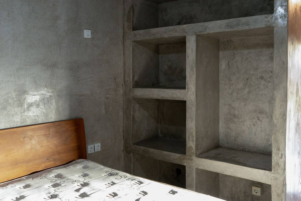 Une chambre avec une tête de lit en bois, un lit recouvert d’une literie à motifs et une étagère en béton adossée au mur.