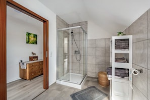 Un bagno moderno con cabina doccia in vetro, un settimino in legno, un porta asciugamani bianco e un quadro appeso al muro.
