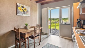 Un coin repas avec une table et des chaises en bois, un tableau représentant des tournesols et, derrière une porte vitrée, une vue sur une terrasse extérieure.