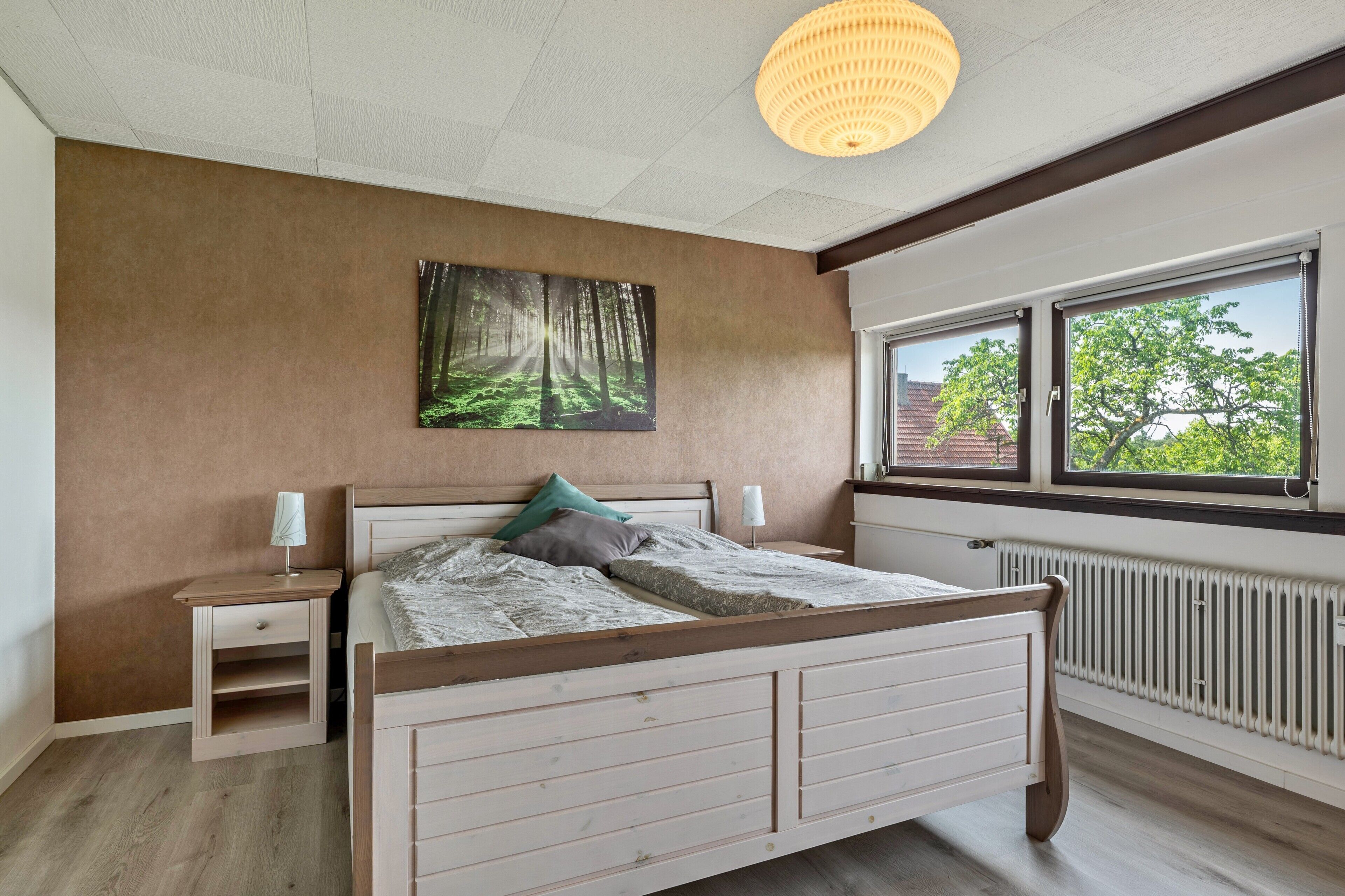 Quarto com cama de madeira, mesas de cabeceira, radiador e janela com vista para árvores.