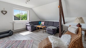 Une pièce de style loft avec un plafond en pente, un canapé d’angle gris, une petite table basse en bois, un fauteuil en osier et une fenêtre donnant sur les arbres.