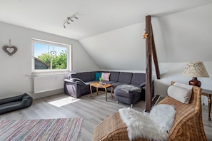 A loft-style room with a sloped ceiling, a gray sectional sofa, a small wooden coffee table, a wicker chair, and a window with a view of trees.
