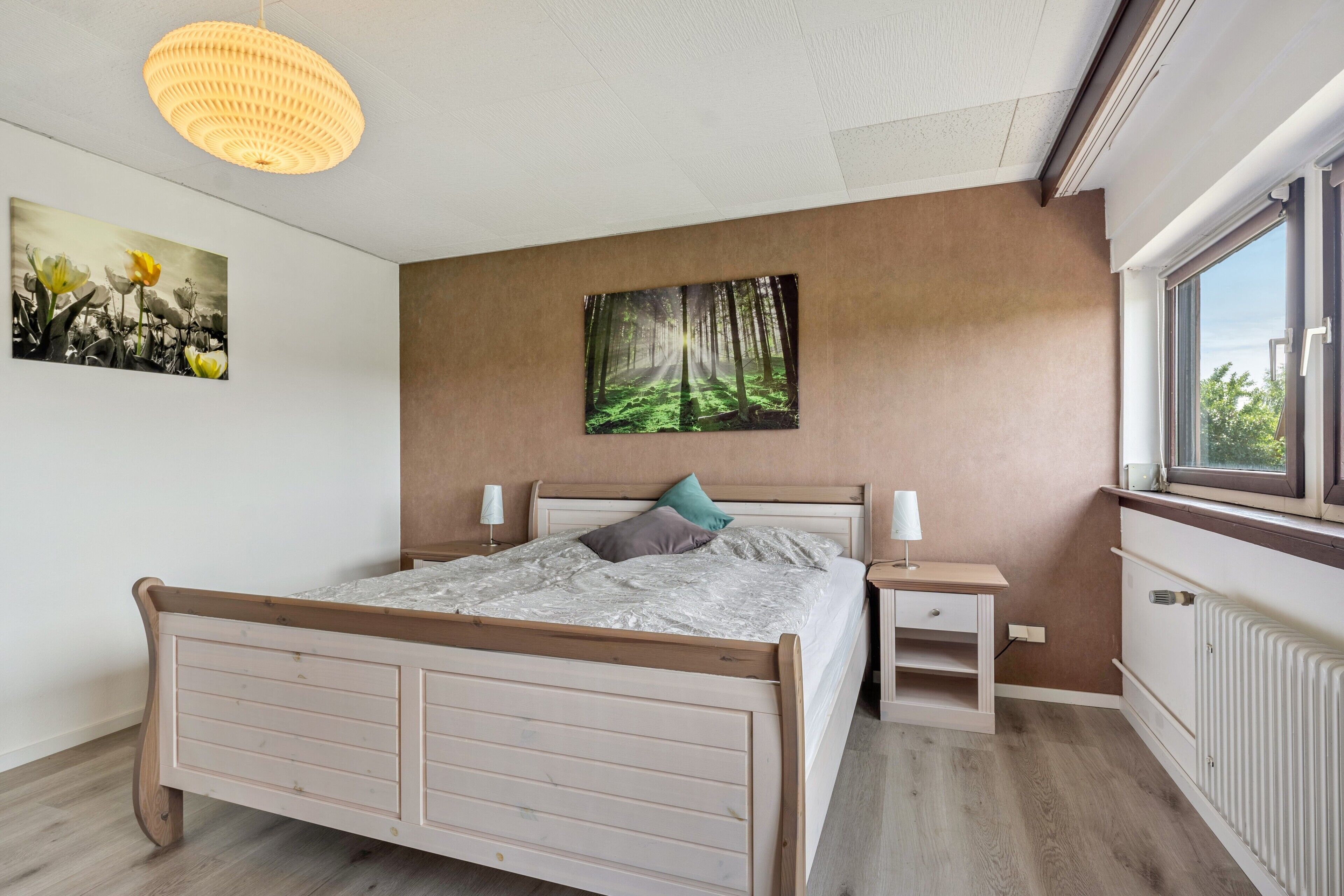 A bedroom with a wooden bed, bedside tables, a wall-mounted painting, and a window with a view.
