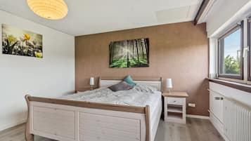 A bedroom with a wooden bed, bedside tables, a wall-mounted painting, and a window with a view.